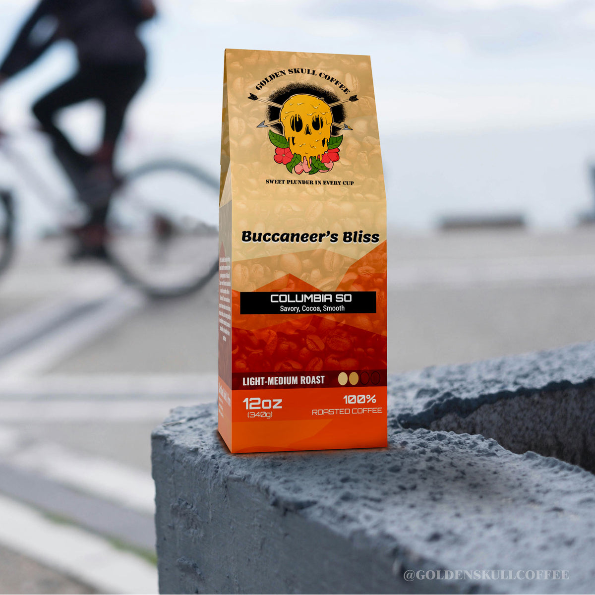 Coffee bag labeled 'Buccaneer's Bliss' by Golden Skull Coffee on a concrete surface with a blurred background of a cyclist.