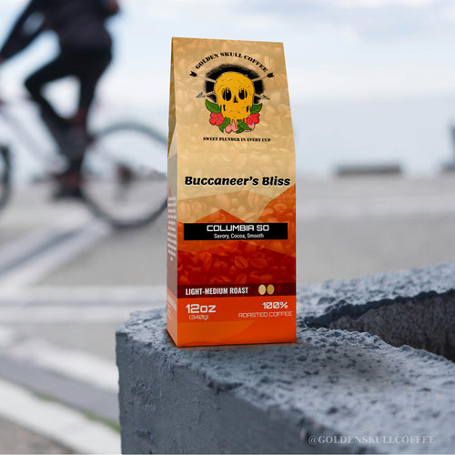 Coffee bag labeled 'Buccaneer's Bliss' by Golden Skull Coffee on a concrete surface with a blurred background of a cyclist.